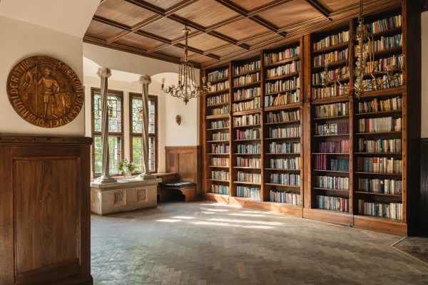 Biblioteka w Zamku Czocha: regały z książkami pod drewnianym kasetonowym stropem, witrażowe okna i rzeźbiony medalion na ścianie
